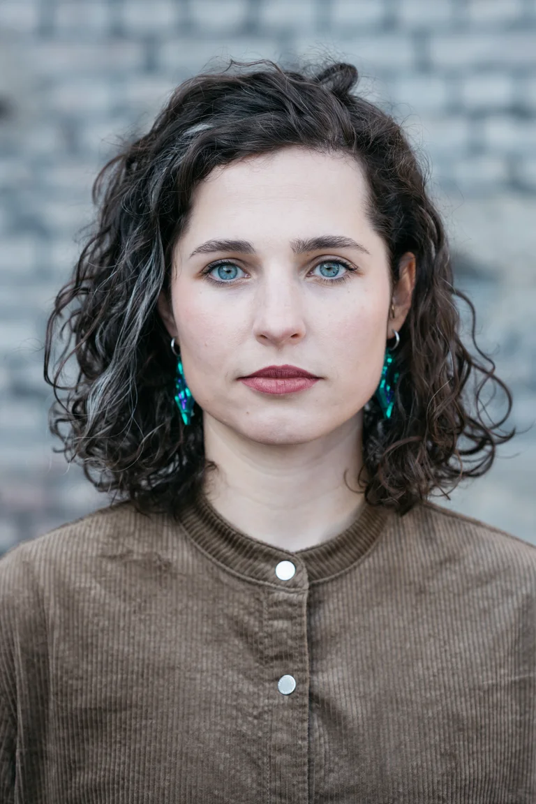 A young woman with her hair down. She stands in front of a gray brick wall and looks directly into the camera.