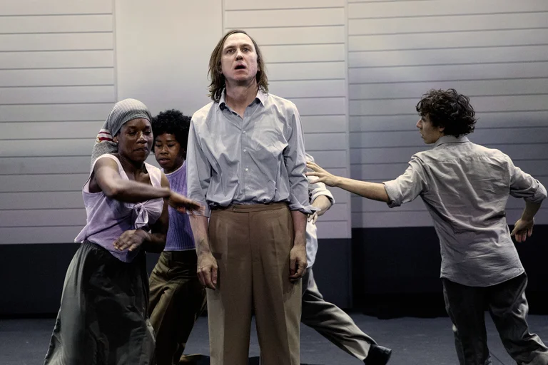 Lars Eidinger stands motionless at the center of a theater stage in front of a light-colored façade, wearing a blank expression. Around him, several performers are captured in motion, including Ida Faho and Sylvie Sanou dressed in purple tops, and Samuel Planas with curly hair, who touches Eidinger from the side.