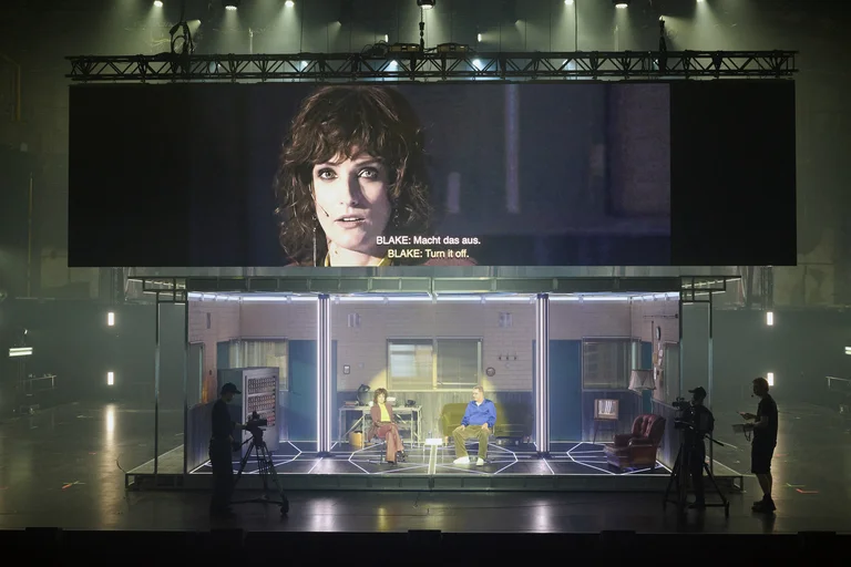 Stage scene with two actors in a room set, above them a large projection with close-up and subtitles.