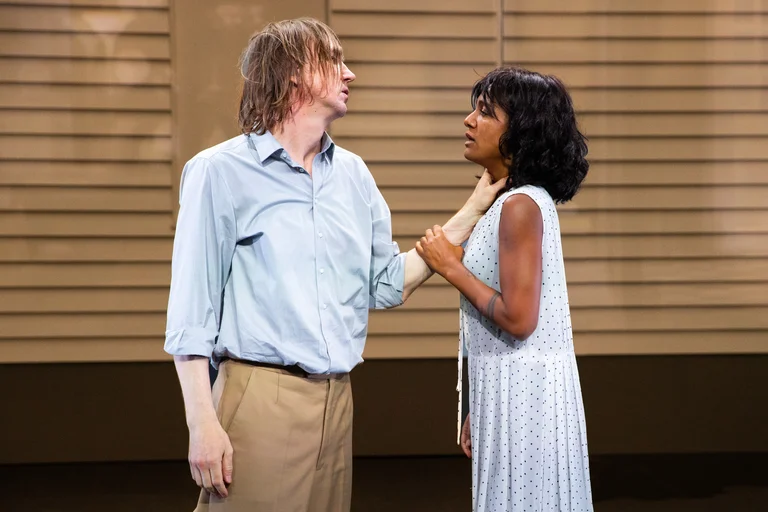 Lars Eidinger and Larissa Sirah Herden stand on a theatre stage in front of a house facade. Eidinger wears a light blue shirt and beige trousers, while Herden is dressed in a white sleeveless dress with small dots. Eidinger holds her by the neck with one hand, while she calmly looks at him and touches his wrist with her hand.