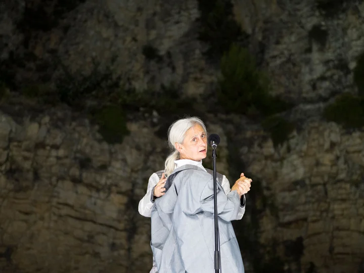 Die Choreografin und Tänzerin Anne Teresa De Keersmaeker dreht sich zur Kamera, vor ihr ein Mikrofon. Hinter ihr steht eine Person, deren Arme zu sehen sind. Sie halten De Keersmaeker an Schulter und der rechten Hand.