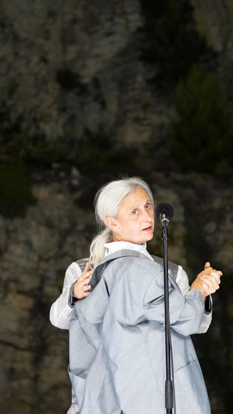 Die Choreografin und Tänzerin Anne Teresa De Keersmaeker dreht sich zur Kamera, vor ihr ein Mikrofon. Hinter ihr steht eine Person, deren Arme zu sehen sind. Sie halten De Keersmaeker an Schulter und der rechten Hand.