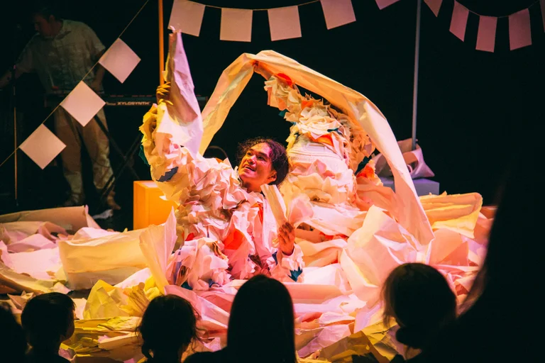 Takeshi Matsumoto sits on a stage in a costume made of crumpled paper, surrounded by large sheets of paper that he holds with outstretched arms. In the background, Makiko Aoyama can be seen wearing a similar costume, partially obscured by the paper sheets. Paper garlands hang above the stage. In the foreground, the silhouettes of audience members are visible as they watch the performance.