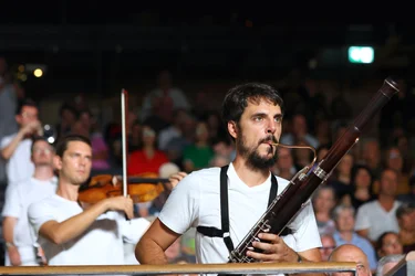 A bassoonist from the ensemble Les Apaches ! performs among the audience. He is wearing a white T-shirt and holds his instrument in front of him. A violinist is playing in the background, with the audience visible behind him.