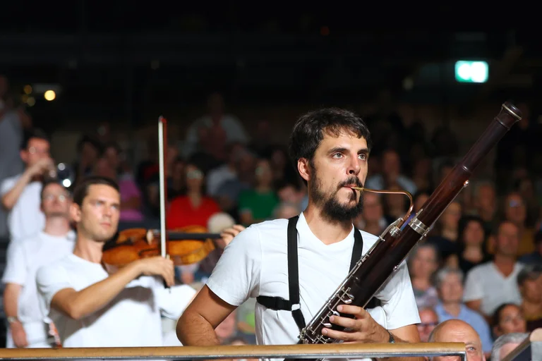 A bassoonist from the ensemble Les Apaches ! performs among the audience. He is wearing a white T-shirt and holds his instrument in front of him. A violinist is playing in the background, with the audience visible behind him.