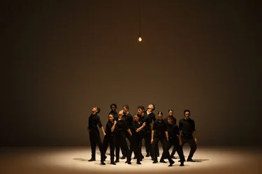 Group of performers in black clothing stand close together under a single light bulb on an empty stage.