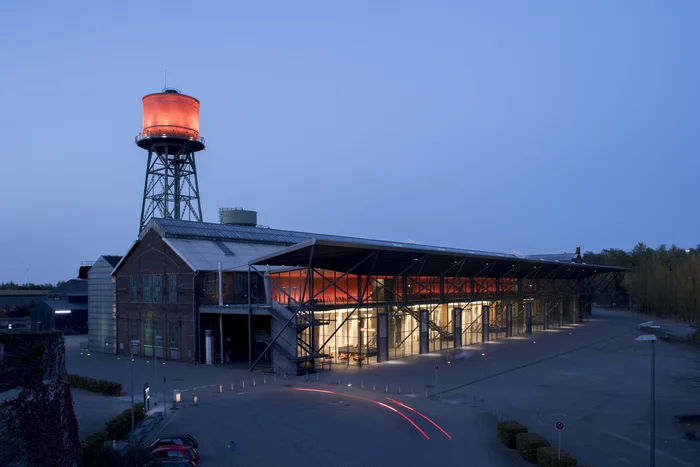 Die Jahrhunderthalle Bochum bei Nacht, beleuchtet durch ein markantes rotes Licht am Wasserturm und den Eingangsbereich, mit Spuren von Autolichtern im Vordergrund.
