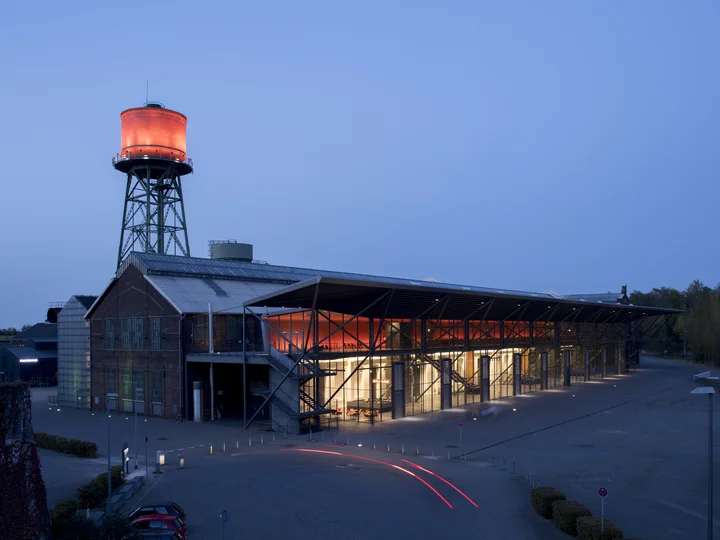 The Jahrhunderthalle Bochum at night, illuminated by striking red light on the water tower and entrance area, with car light trails in the foreground.
