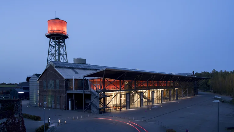 The Jahrhunderthalle Bochum at night, illuminated by striking red light on the water tower and entrance area, with car light trails in the foreground.