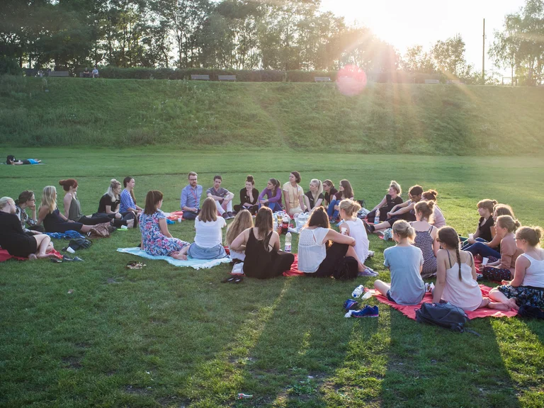 Junge Menschen sitzen in einem Kreis auf einer Wiese bei strahlendem Sonnenschein und unterhalten sich entspannt.