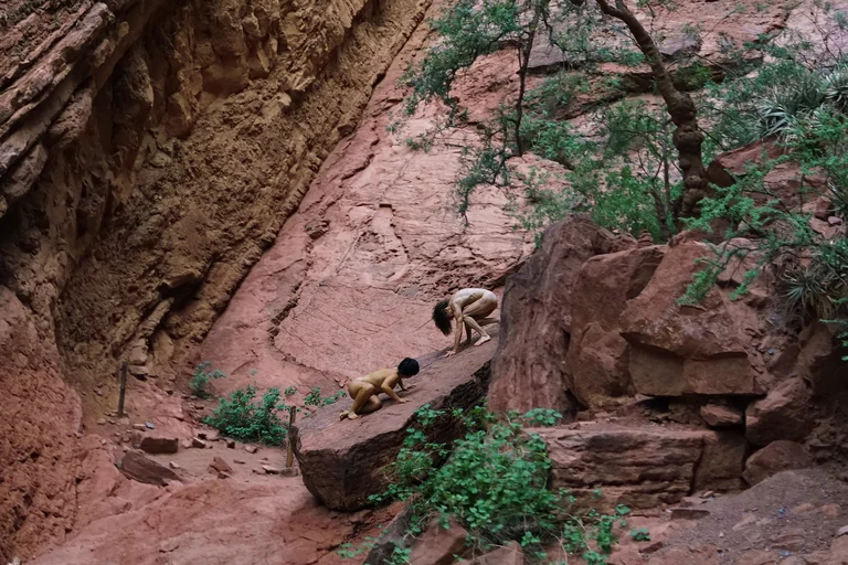 A canyon with red rock formations and lush green vegetation. Two people can be seen in the center of the image, bending over a rock.