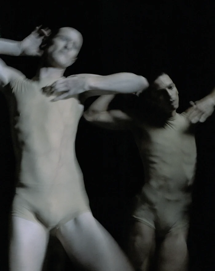 Two dancers in skin-toned costumes move powerfully and expressively against a black background. The blurred motion enhances the sense of intensity and fluid movement.