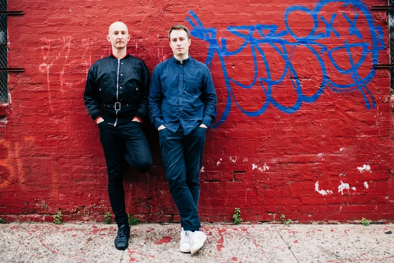Two men posing in front of a red wall with grafitti