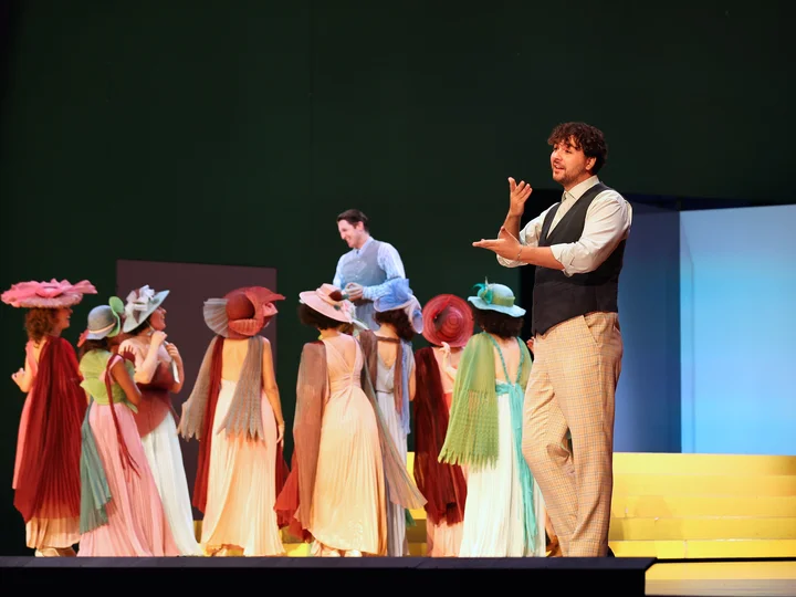 A sign language interpreter in a checkered suit and vest translates on stage, while a group of performers in pastel-colored dresses and striking hats face away from him. Another actor in a blue costume stands in the background.
