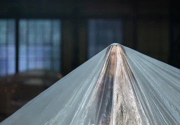 A person stands beneath a large sheet of translucent plastic stretched tent-like over their head and body. Large windows are visible in the dark, blurred background.