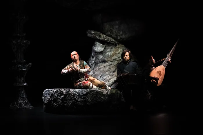 Eine Performer*in liegt auf einem Felsen, den Oberkörper aufgerichtet, einen Arm ausdrucksstark in die Höhe gestreckt. Rechts daneben sitzt ein Musiker mit einem Bandoneon und schaut aufmerksam zur Szene.