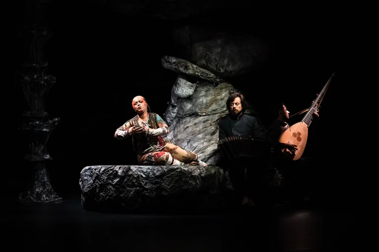 A performer reclines on a rock, torso raised, with one arm expressively reaching upward. To the right, a musician with a bandoneon watches the scene attentively.