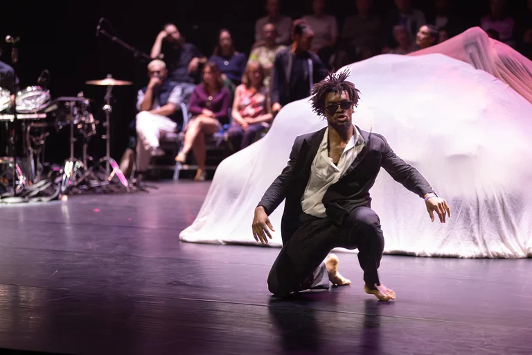 A dancer in a black suit and white shirt is kneeling barefoot on the stage. He is wearing sunglasses.
