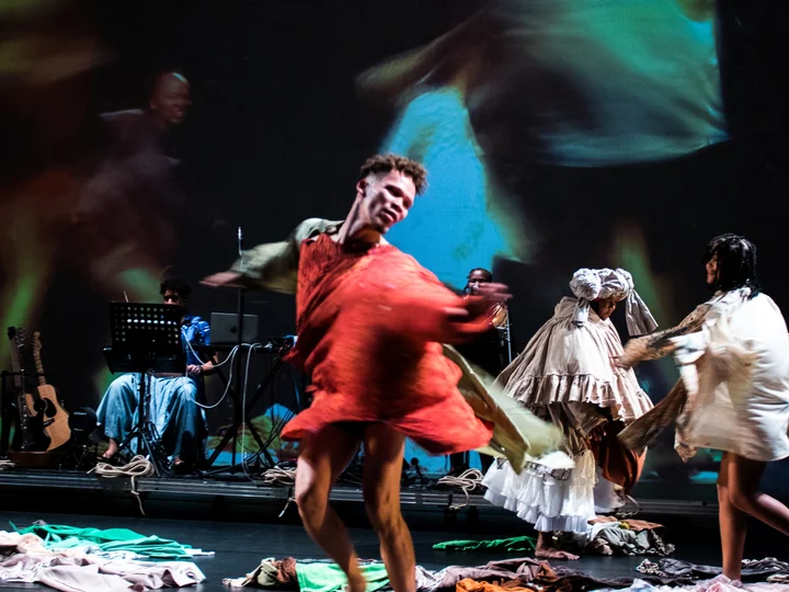 A dancer in a flowing red costume twirls dynamically across the stage, while other performers in layered garments move in the background. Colorful fabrics are scattered across the floor, and blurred, moving projections are visible on the backdrop.