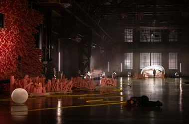 Wide view of a dark industrial hall featuring a stage installation with numerous pink foam elements, lights, and yellow cables across the floor. A person lies on the floor in the foreground, while two others stand in the hall. An illuminated inflatable structure is visible in the background.
