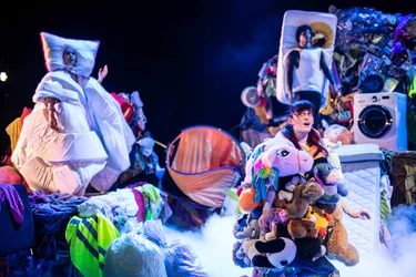 Bühnenszene mit mehreren Performer:innen zwischen großen Haufen aus Kleidung, Haushaltsgegenständen und Matratzen; im Vordergrund steht eine Person in einem Kostüm, das aus vielen Stofftieren besteht.