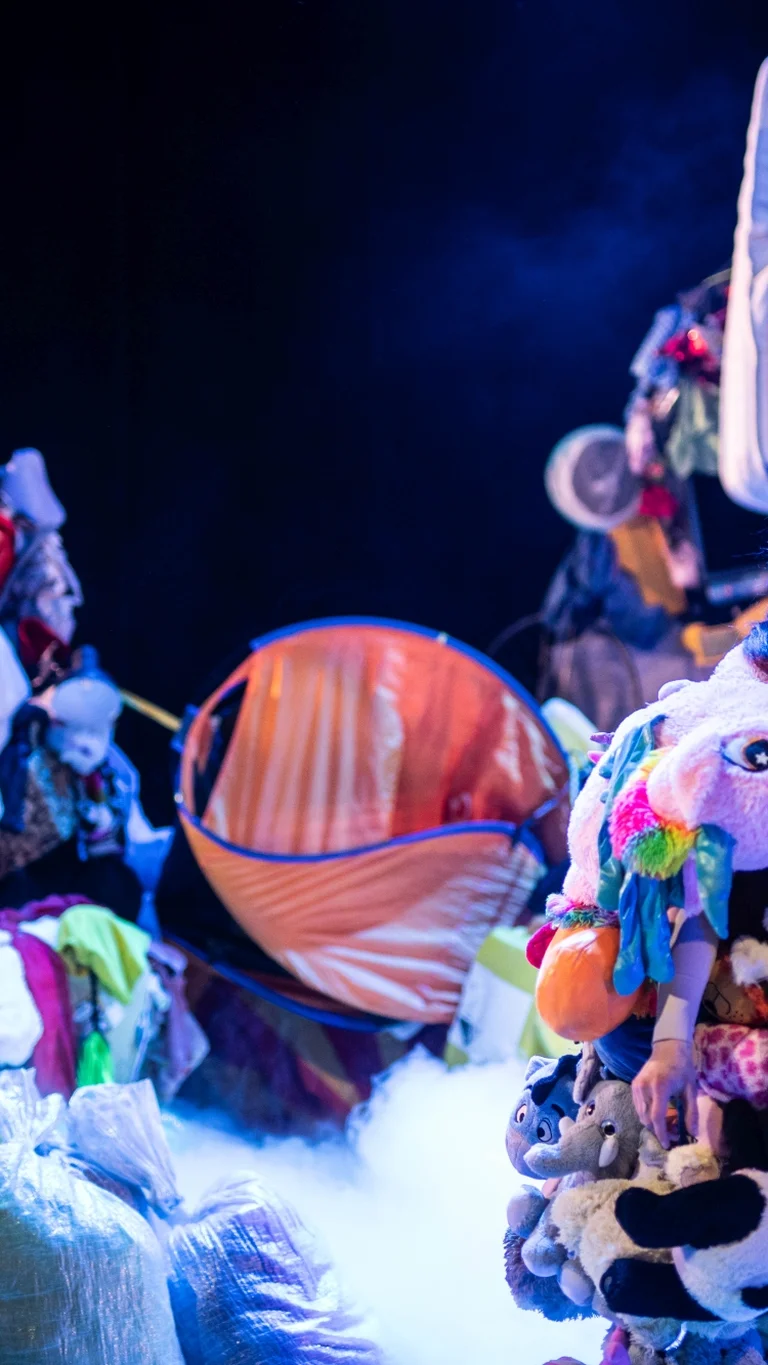 Bühnenszene mit mehreren Performer:innen zwischen großen Haufen aus Kleidung, Haushaltsgegenständen und Matratzen; im Vordergrund steht eine Person in einem Kostüm, das aus vielen Stofftieren besteht.