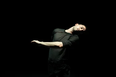 A dancer in black tilts his torso to the side and extends one arm; the scene is minimally lit against a black background.