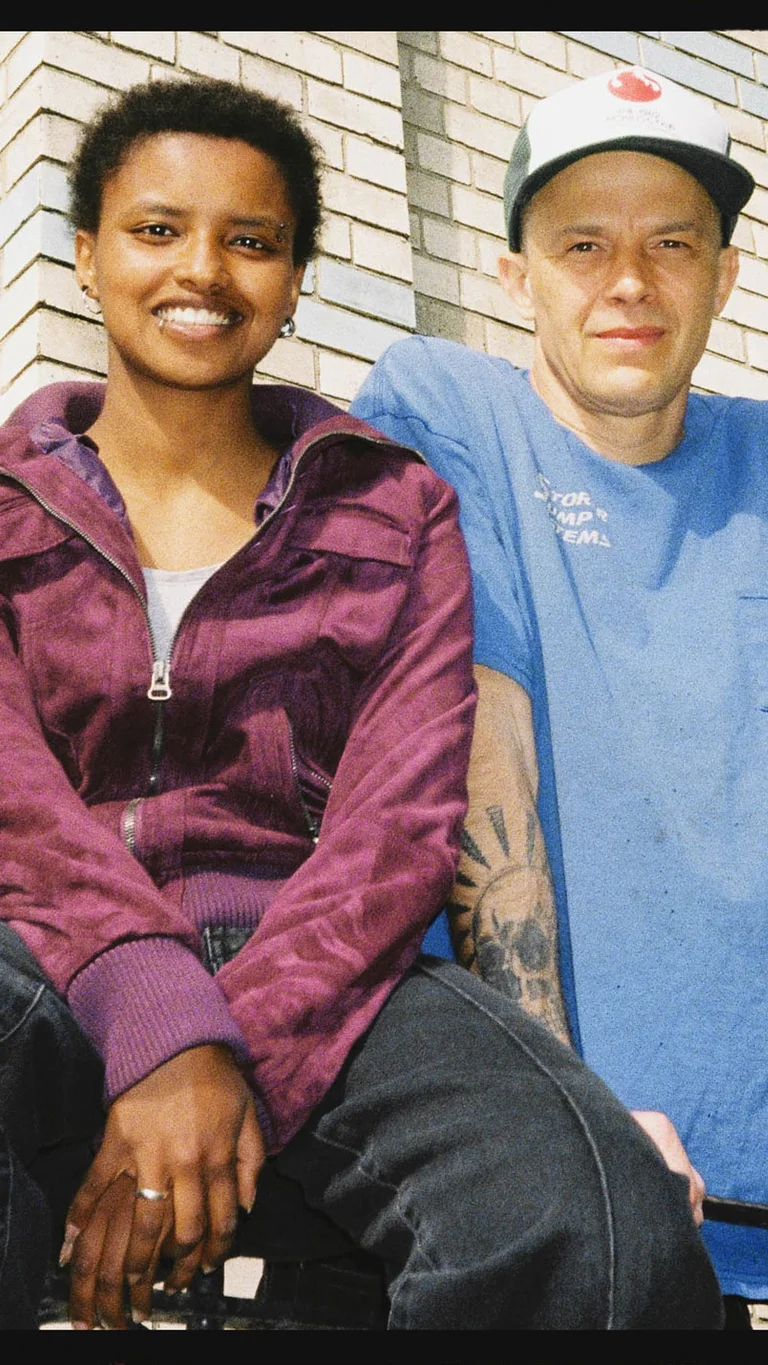 Meskerem Mees and Carlos Garbin sit next to each other on a railing in front of a house facade with windows, looking at the camera.