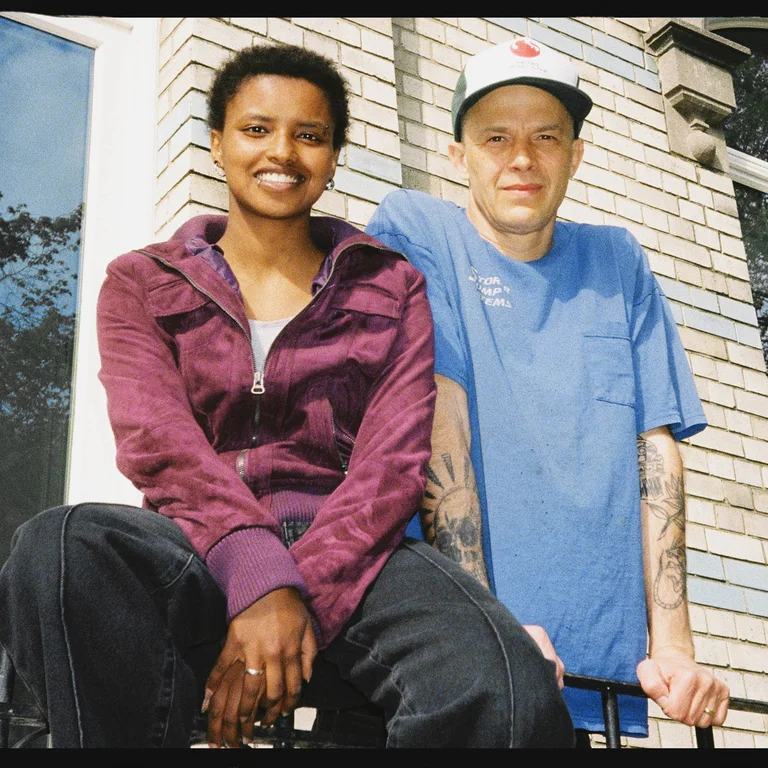 Meskerem Mees und Carlos Garbin sitzen nebeneinander auf einem Geländer vor einer Hausfassade mit Fenstern und schauen in die Kamera.
