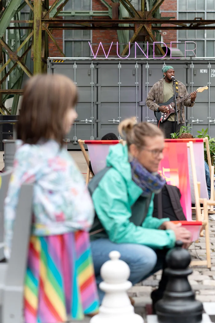 Joël-João spielt E-Gitarre und singt auf einer Bühne vor dem Schriftzug „WUNDER“. Im Vordergrund sind unscharf Kinder und Erwachsene zu sehen.