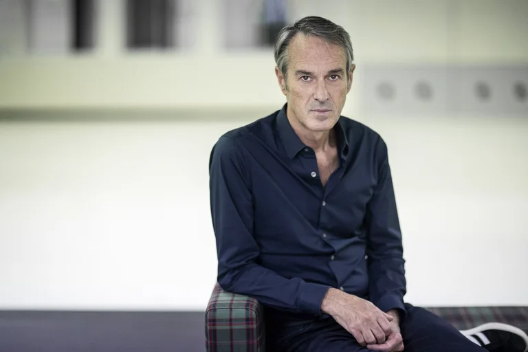 Ivo Van Hove, artistic director of the Ruhrtriennale 2024-2026, sits in a checked armchair. He is wearing a dark blue shirt and looking directly into the camera.