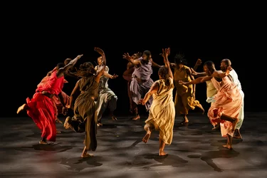 Eine Gruppe von Tänzer:innen bewegt sich barfuß in einer dynamischen Choreografie über eine dunkle Bühne. Sie tragen weit fließende Kostüme in kräftigen Rot-, Orange-, Gelb-, Braun- und Grautönen. Vor schwarzem Hintergrund werden ihre Bewegungen und erhobenen Arme beleuchtet.