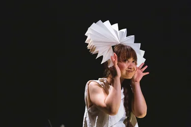Die Performerin Makiko Aoyama mit einem kunstvollen Kopfschmuck aus Papier lächelt und macht eine spielerische Geste mit ihren Händen.